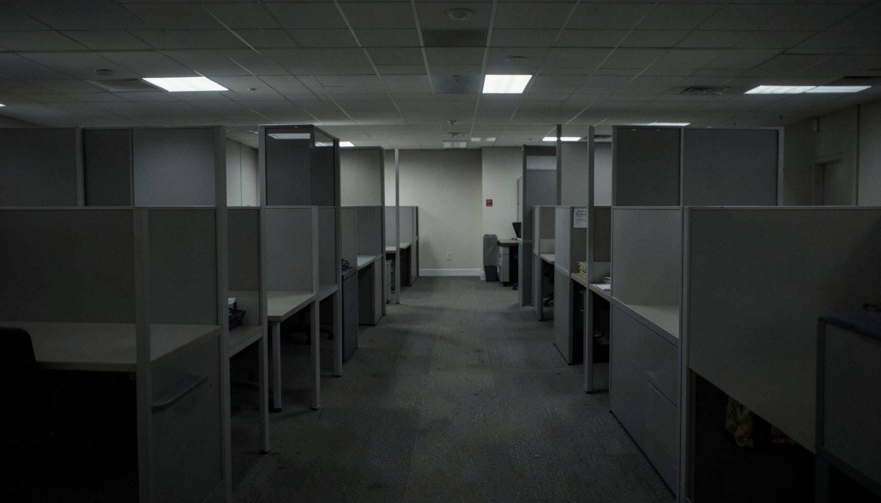 A dim office corridor with empty desks and grey cubicles in a quiet subdued mood