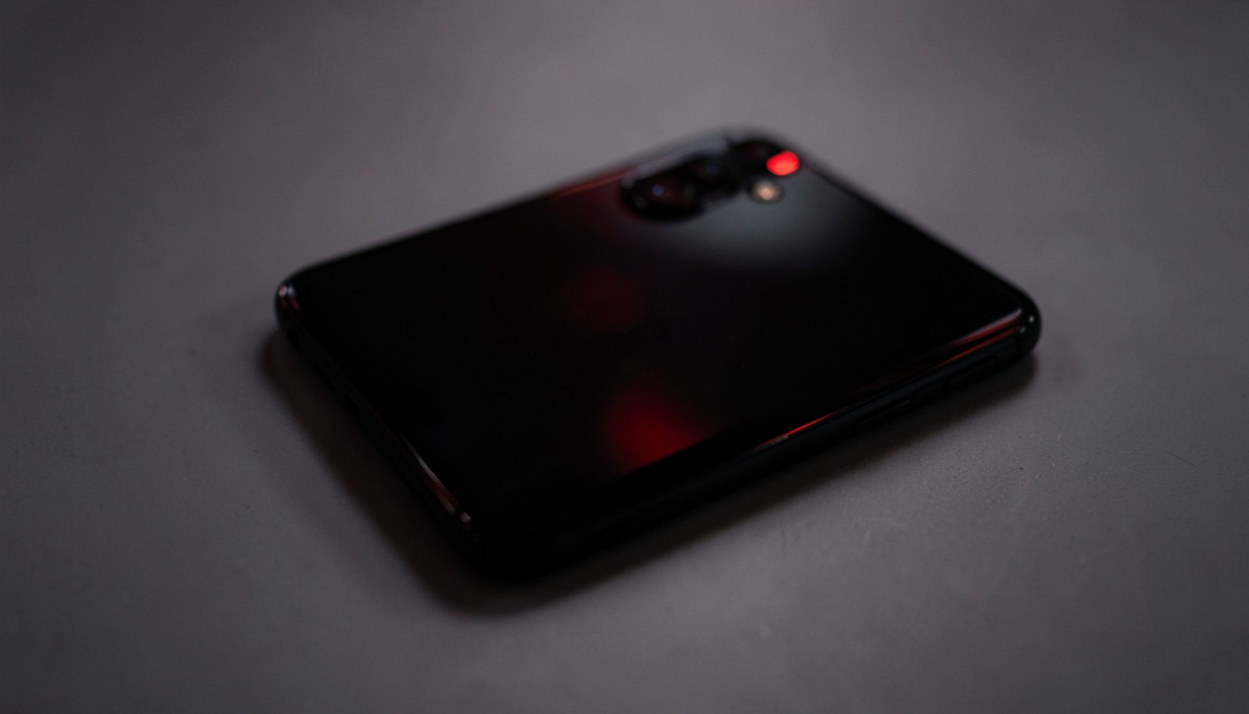 A dark smartphone with a glossy back panel resting on a plain grey desk, moody low-key lighting with a red accent reflection